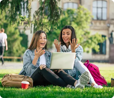 Two students sitting on a campus lawn using a laptop together.