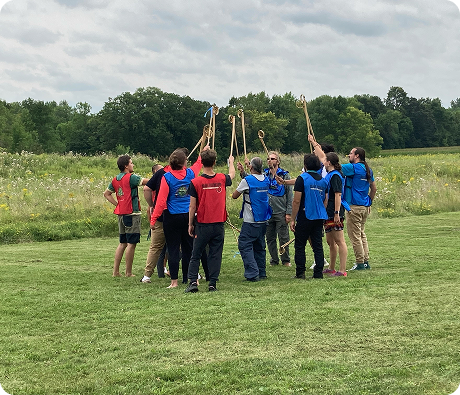 Group of people gathered outdoors in a grassy meadow, participating in a community activity with wooden poles.